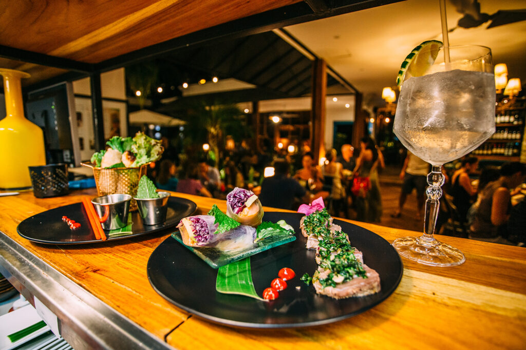 A close-up shot of fresh spring rolls and seared tuna appetizers plated on dark dishes, served alongside a iced cocktail with a lime wedge on a wooden bar top at a lively Ojochal, Costa Rica restaurant.