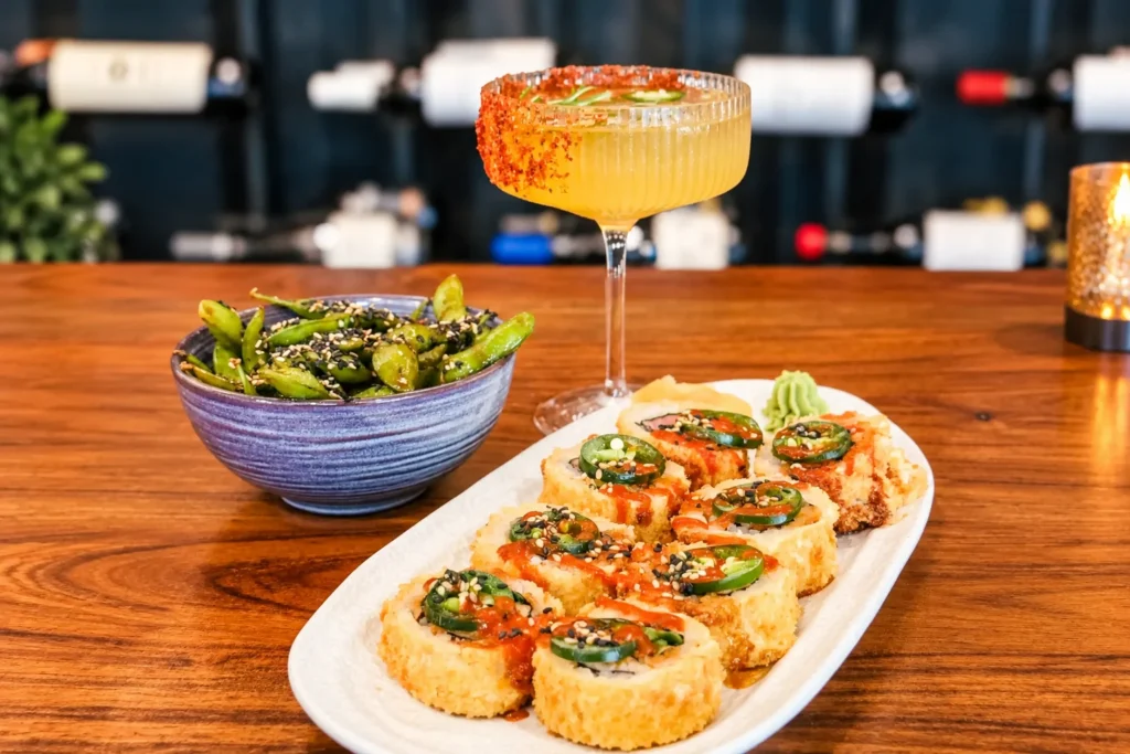 A platter of crisp specialty sushi rolls topped with sliced jalapeños and spicy sauce, served next to a bowl of seasoned edamame and a chili-rimmed cocktail on a polished wooden table.