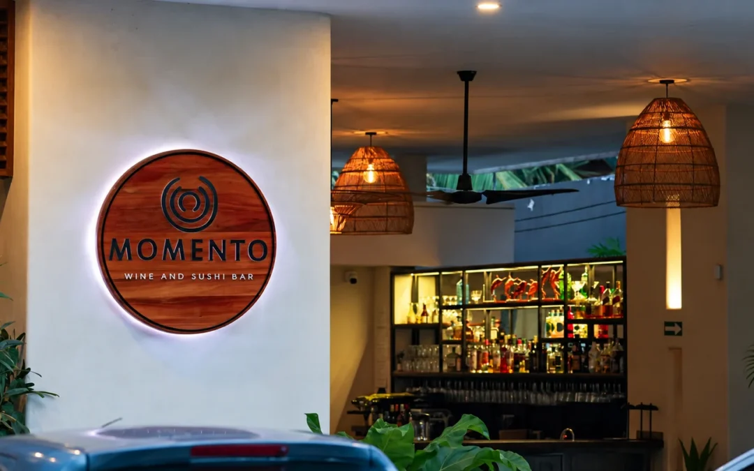A illuminated wooden circular sign for Momento Wine and Sushi Bar mounted on a white exterior wall next to an open-air dining space featuring woven basket pendant lights and a fully stocked bar.
