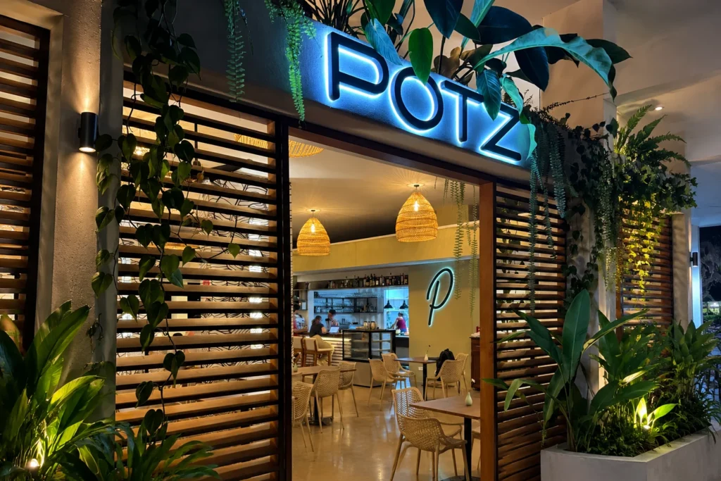 A blue illuminated Potz sign glowing above a modern restaurant entrance decorated with wooden slatted walls, lush tropical plants, and warm woven basket light fixtures inside.