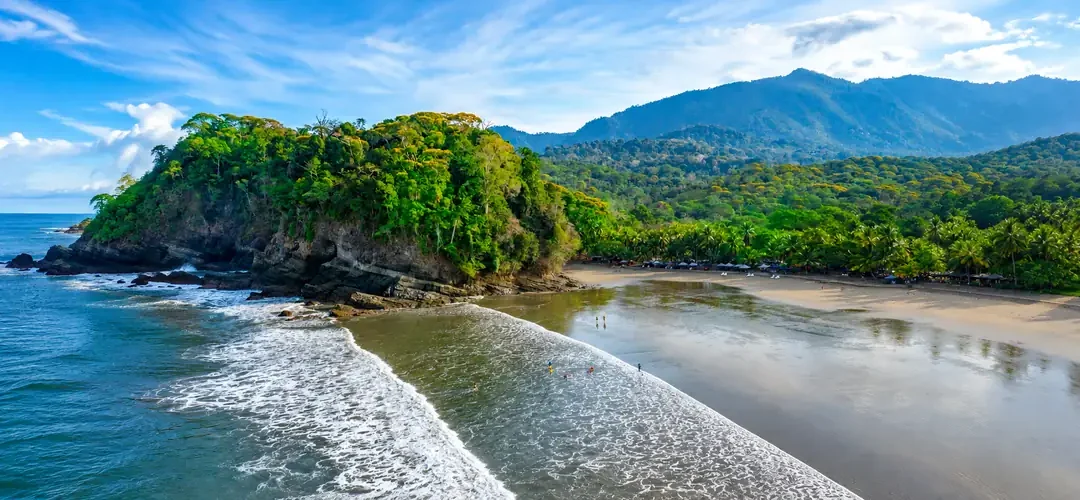 Aerial view of a jungle-covered headland and dark sand beach backed by palm trees and mountains on Costa Rica’s south Pacific coast